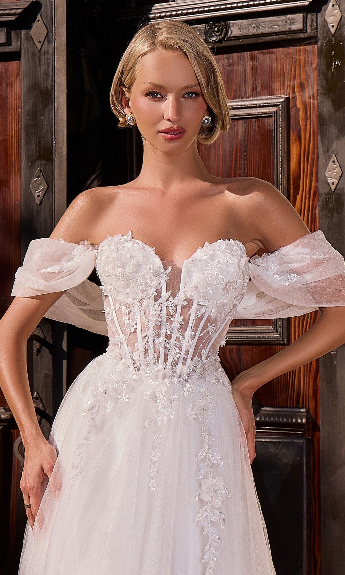 A model poses gracefully in front of a wooden door, wearing the Ladivine CD2426W long formal dress: a white off-the-shoulder bridal gown featuring a sheer corset bodice, floral embroidery, and puff sleeves.